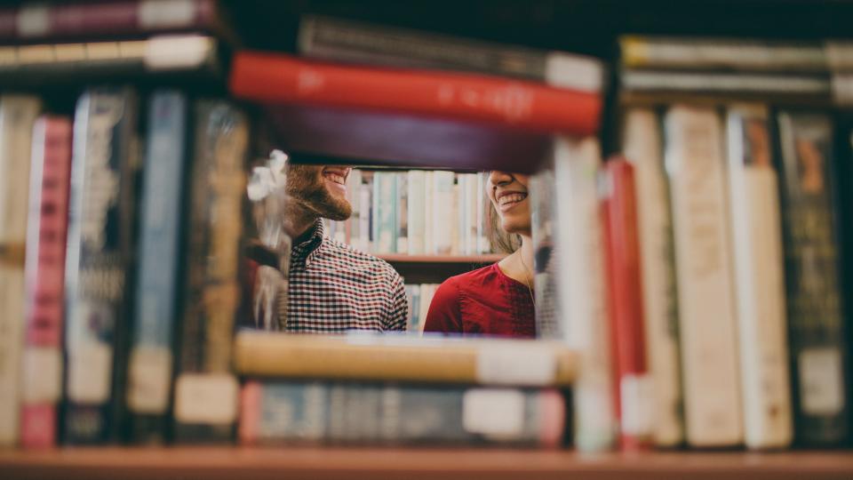 Two students smiling through a gap in library bookshelves